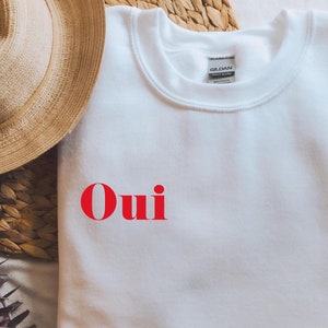 Puede incluir: Sudadera blanca con la palabra "Oui" en letras rojas.