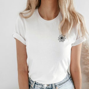 May include: White t-shirt with a small black daisy graphic on the chest. The shirt has short sleeves and a crew neck. The model is wearing light blue jeans.
