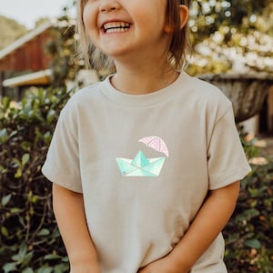 Może przedstawiać: Jasnobeżowa koszulka z jasnoniebieską łódką z papieru, na której znajduje się różowy parasol w białe kropki. Koszulkę nosi dziecko.