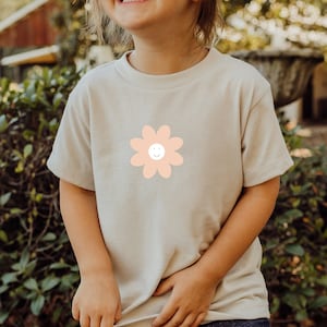 Könnte beinhalten: Ein hellbeiges T-Shirt mit einem pfirsichfarbenen Blumenmotiv. Die Blume hat ein lächelndes Gesicht.