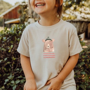 Peut inclure: T-shirt beige avec une illustration à l'aquarelle d'un ours portant un bonnet vert. En dessous de l'ours, trois rayures horizontales rouges. Le t-shirt est à col rond et à manches courtes.