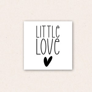 May include: A square white card with the words "LITTLE LOVE" in black script, with a black heart below the word "LOVE". The card is set against a light beige background.