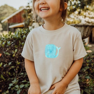 Pode incluir: Uma camiseta bege com uma ilustração de baleia em aquarela azul claro. A baleia tem um rosto sorridente e uma cauda curva. A camiseta é usada por uma criança. O fundo está desfocado, mostrando vegetação e uma estrutura de madeira.