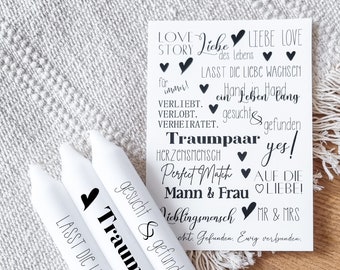 May include: White candles with black text that reads "Traumpaar" and "Lasst die Liebe wachsen". The candles are on a gray and brown surface. The text on the candles is in German and translates to "Dream Couple" and "Let love grow".