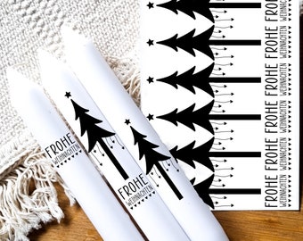 May include: Three white candles with black Christmas tree designs and the text "Frohe Weihnachten" printed on them. The candles are on a wooden surface with a white sheet in the background. A sheet of paper with black Christmas tree designs and the text "Frohe Weihnachten" printed on it is also visible.
