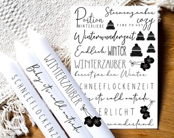 Puede incluir: Tres velas blancas con texto negro e ilustraciones. El texto en las velas dice "Winterzauber", "Baby it's cold outside" y "Schneeflockenzeit". Las ilustraciones incluyen copos de nieve y guantes.