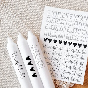 Op de afbeelding: Drie witte kaarsen met zwarte tekst. De kaarsen zijn gelabeld met "Wunschlicht" en "Glückslicht". De tekst is in het Duits. Er zijn zwarte harten op de kaarsen.
