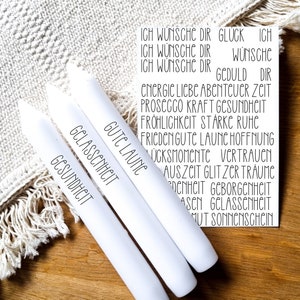 May include: Three white candles with black text. The text on the candles reads "Gute Laune", "Glasschmeckt", and "Gesundheit". The candles are on a wooden surface with a white fuzzy blanket in the background. The text on the top of the image reads "Ich wünsche dir Glück", "Ich wünsche dir", "Ich wünsche dir Geduld", "Energie Liebe Abenteuer Zeit", "Prosecco Kraft Gesundheit", "Fröhlichkeit Stärke Ruhe", "Frieden Gute Laune Hoffnung", "Glücksmomente Vertrauen", "Auszeit Gltiz der Träume", "Zärtlichkeit Geborgenheit", "Aschen Glasschheit", "Mut Sonnenschein".