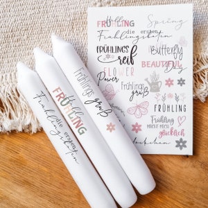 May include: Three white candles with the text "FRÜHLING" in pink, green, and white lettering. The candles are decorated with a pink flower. The background is a white card with the text "FRÜHLING" in various colors and designs.