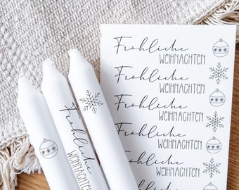May include: Three white candles with black text that reads "Frohliche Weihnachten" and a snowflake or ornament design. The candles are on a white textured surface.