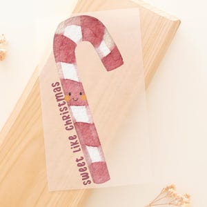 Könnte beinhalten: Ein rechteckiges, transparentes Blatt mit einer aquarellartigen Illustration einer rot-weiß gestreiften Zuckerstange mit einem kleinen lächelnden Gesicht und rosa Wangen. Der Text "sweet like Christmas" ist vertikal in Braun neben der Zuckerstange gedruckt.