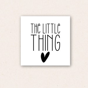 May include: A square white card with the words "THE LITTLE THING" in black, stylized font. Below the word "THING" is a black heart. The card is on a light beige background.