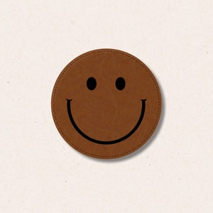 Op de afbeelding: Een bruine leren smiley-patch met zwarte stiksels rond de randen en een zwart smiley-ontwerp.