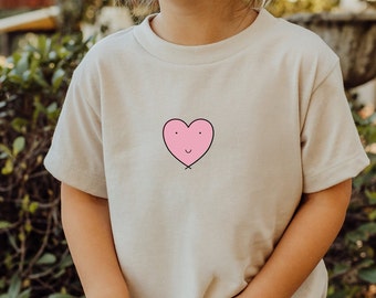 May include: A child wearing a light beige t-shirt with a pink heart graphic. The heart has a smiling face.