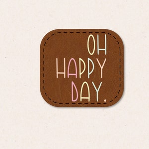 Può includere: Un cerotto in pelle marrone con un bordo cucito e il testo "OH HAPPY DAY." in lettere colorate.