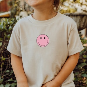 Peut inclure: Un enfant portant un t-shirt beige clair avec un smiley rose.