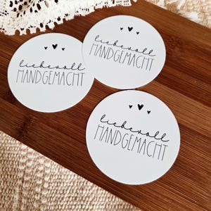 May include: Three white, round tags with the German text "liebevoll HANDGEMACHT" in black script and block letters. Each tag has a small heart-shaped hole at the top and two small black heart illustrations. The tags are on a wooden surface.