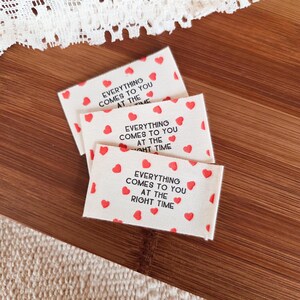 Könnte beinhalten: Drei rechteckige Stoffetiketten mit dem Text "EVERYTHING COMES TO YOU AT THE RIGHT TIME" in Schwarz, umgeben von kleinen roten Herzen. Die Etiketten liegen auf einer Holzoberfläche.