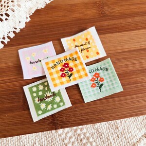 May include: A collection of five fabric labels with floral designs and the words "HAND MADE" and "handmade". The labels feature various colors, including pink, yellow, and green, with embroidered flowers and gingham patterns. The labels are square and appear to be for sewing projects.