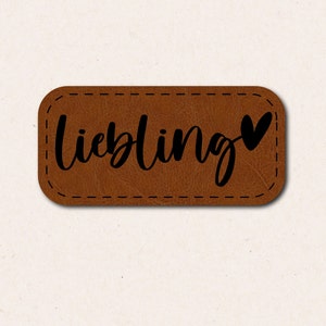 May include: A brown leather patch with a stitched border and the word "Liebling" in black script with a small heart at the end.