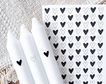 May include: Three white candles with black heart designs on them. The candles are next to a sheet of paper with a pattern of black and white hearts.