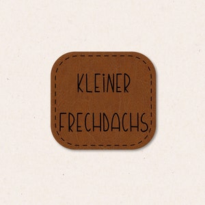 Può includere: Un cerotto in pelle marrone con il testo "KLEINER FRECHDACHS" in nero.
