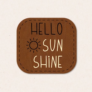 Pode incluir: Um remendo de couro marrom com uma borda costurada e o texto "HELLO SUNSHINE" em letras pretas e brancas. Um pequeno gráfico de sol está acima da palavra "SUN".