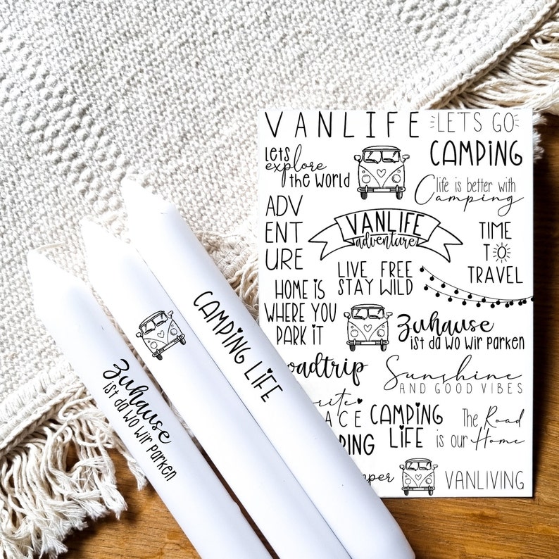 May include: Three white candles with black text and a graphic of a vintage camper van. The text on the candles reads "Camping Life" and "Zuhause ist da wo wir parken".