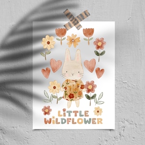 Puede incluir: Una ilustración de acuarela de un conejo blanco con un vestido floral y sosteniendo una flor. El conejo está rodeado de corazones, flores y el texto "little wildflower".