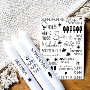 May include: Three white candles with black text and images. The candles are labelled "Hallo Sommer", "SUMM SUMM SUMM", and "SOMMERMOMENTE". The image also includes a sheet of stickers with summer-themed text and images, such as "Aloha Vibes", "Melonlove", "Sommerlicht Auszeit", and "Sommergruss".