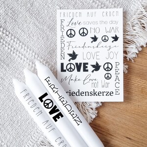 May include: Three white candles with black text and peace signs. The text on the candles reads "Frieden", "Love", and "Frieden auf Erde".