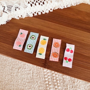 May include: Six fabric hair clips with fruit designs. The clips feature patterns of pink peaches, green avocados, yellow pineapples, orange slices, and red cherries. Each clip is rectangular and has a checkered background.