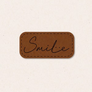 Puede incluir: Un parche de cuero marrón con un borde cosido y la palabra "Smile..." escrita en negro.