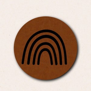 May include: A round, brown leather coaster with a black rainbow design. The rainbow consists of five curved lines, creating an arch shape. The coaster has a textured surface and a simple, modern aesthetic.