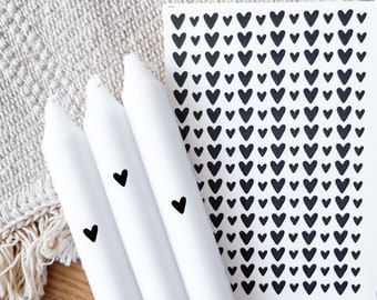 May include: Three white taper candles with black heart designs. A sheet of paper with a repeating pattern of black hearts is in the background.