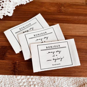 Puede incluir: Tres etiquetas rectangulares de tela blanca con la palabra "BONJOUR" en la parte superior y la frase "every day is a new beginning" en escritura manuscrita. Las etiquetas tienen texto negro y un borde negro. Las palabras "hand made" están en la parte inferior.