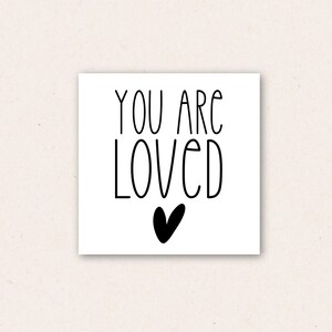May include: A white card with black text that reads "You are loved" with a black heart below the text.