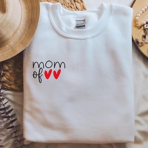 Puede incluir: Sudadera blanca con el texto "mom of" y dos corazones rojos.