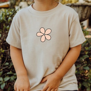 Può includere: Una maglietta beige chiaro con una grafica di un fiore rosa.