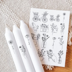May include: Four white candles with black line drawings of flowers and words like "breathe", "love", "smile", and "hope". The candles are next to a sheet of paper with black line drawings of flowers, a bicycle, a camera, and a jar.