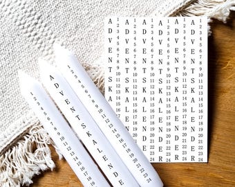 May include: Three white candles with the words "Adventkalender" printed on them. The candles are numbered 1 through 24. The candles are on a wooden surface with a white crocheted blanket in the background.