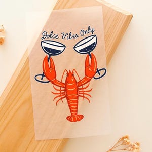 Puede incluir: Una lámina transparente con una ilustración de una langosta roja sosteniendo dos copas de vino. El texto "Dolce Vibes Only" está escrito encima de la langosta. La lámina está colocada sobre una superficie de madera.