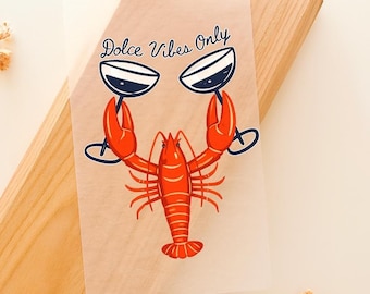 May include: A transparent sheet with a red lobster illustration holding two wine glasses. The text "Dolce Vibes Only" is written in a cursive font above the lobster. The sheet is placed on a wooden surface.