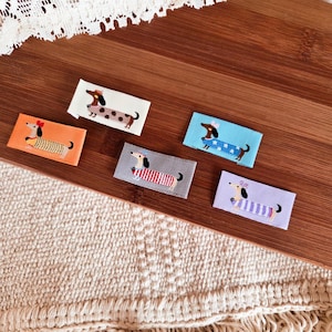 Puede incluir: Una colección de parches bordados de perros salchicha en varios colores. Cada parche presenta un diseño diferente de perro salchicha, incluyendo lunares, rayas y atuendos. Los parches son rectangulares y están sobre una superficie de madera.