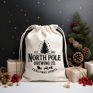 Könnte beinhalten: Ein cremefarbener Kordelzugbeutel mit dem Text "North Pole Brewing Co. Christmas Spirits" in Schwarz. Das Design umfasst einen Weihnachtsbaum, einen Schlitten und Rentiere. Der Beutel ist von verpackten Geschenken, Tannenzapfen und Beeren umgeben.