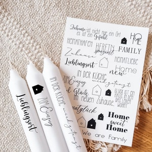May include: Three white candles with black text. The text on the candles reads "Lieblingsort", "Zum Einzug", and "In der K&uuml;che wird getanzt". The candles are on a white and brown textured surface. A white card with black text is behind the candles. The text on the card reads "Zuhause ist nicht nur ein Ort, es ist ein Gef&uuml;hl", "Heimathafen", "Herzen", "Family", "Zuhause", "Lieblingsort", "In der K&uuml;che", "Zum Einzug", "el Gl&uuml;ck", "im neuen Zuhause", "natgl&uuml;ck", "Home sweet Home", "We are Family", "Heimatliebe", "Home", "new", "getanzt", "Gl&uuml;ck ist ein Haus in dem das Leben wohnt".