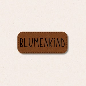 Könnte beinhalten: Ein braunes Leder-Etikett mit dem Text "BLUMENKIND" in schwarzen Buchstaben.