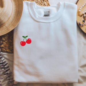 Puede incluir: Sudadera blanca de cuello redondo con un pequeño gráfico de cereza roja en el lado izquierdo del pecho.