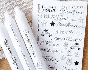 May include: Three white candles with black lettering that reads "Merry Christmas", "Christmastime", and "Christmas lights". A sheet of white paper with black Christmas-themed stickers is in the background. The stickers include the words "Santa", "Christmas", "Christmas Magic", "Christmastime", "Jingle All The Way", "Christmas Lights", and "Christmas Feeling".