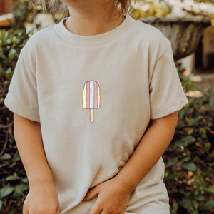 Könnte beinhalten: Ein hellbeiges T-Shirt mit einem Comic-Eis am Stiel in den Farben Pink, Gelb und Blau.